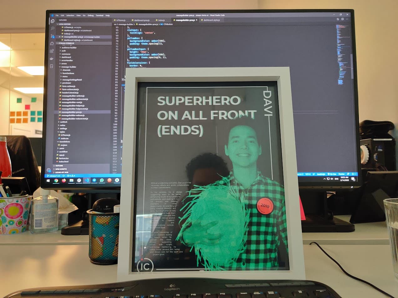 A picture frame of Davi as part of an employee award. The picture frame is on top of a desk with a monitor displaying code on the background